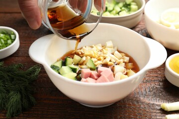 Making okroshka soup. Woman pouring kvass into bowl with different ingredients at wooden table, closeup