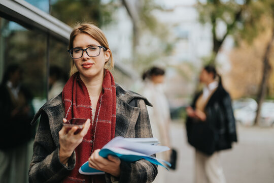 Confident woman standing outdoors with a smart phone and papers, dressed in professional attire. Ideal representation of professional women, business networking, and productivity in an urban setting.