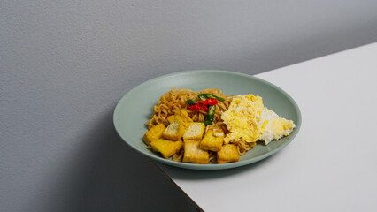 A plate of fried noodles served with egg and fried tofu. Topped with red and green chilies.