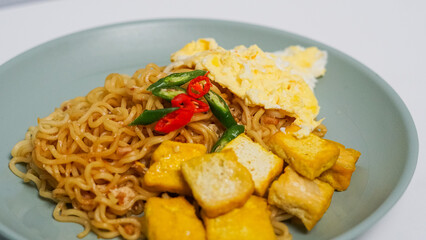 A plate of fried noodles served with egg and fried tofu. Topped with red and green chilies.