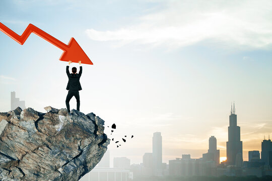 Businessman holding red downward arrow on cliff, symbolizing financial crisis, with city skyline in background, conveying economic collapse.