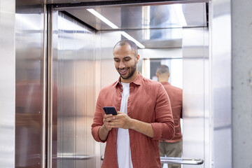 A man is smiling in an elevator while looking at his smartphone, wearing a casual terracotta shirt and a white t-shirt © Liubomir