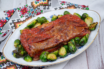 Vegan pork bellly roast made with seitan and starch, served on oval plate along with roasted Brussels sprouts. Wooden table, stylish setting