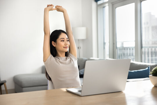 Young Asian woman doing relaxation exercises, stretching her arms upward while sit at workplace desk in front of laptop, smile, take break from work or studying at home office. Rest, work-life balance