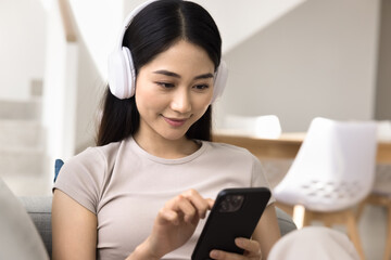 Close up of young Asian woman wear headphones using smartphone sitting comfortably on couch. Female...