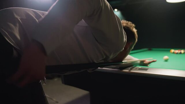 Close-up backshot of player in white shirt leaning over green pool table, gripping cue stick with intense focus. Dark background enhances concentration as he prepares to strike white cue ball
