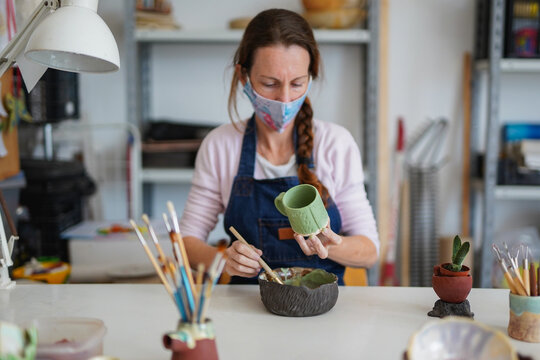 Caucasian woman working with ceramic and paint finished cup while wearing  face mask - Art work studio - Focus on hand
