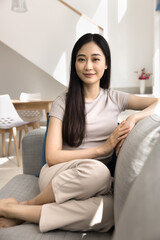 Beautiful young Asian woman dressed casually relaxing seated comfortably on sofa in modern sunny living room, looking at camera, posing, exuding contentment and inner peace. Lifestyle and well-being