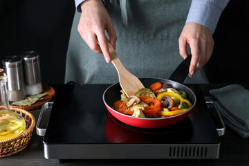 Woman frying vegetables and mushrooms in pan on stove at wooden table, closeup