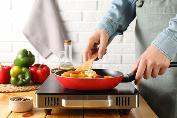 Woman frying vegetables in pan on stove at wooden table indoors, closeup