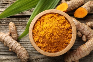 Turmeric powder in bowl, roots and leaves on wooden table, flat lay