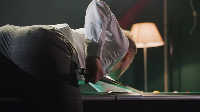 Close-up back shot of billiard player in white shirt and grey trousers leaning over green pool table, gripping cue stick with precision. Overhead lighting casts warm glow as he lines up strategic shot