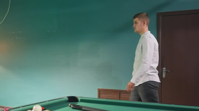 Side shot of young man in white shirt walking into billiard room with modern interior, approaching pool table, and picking up cue stick. Green pool tables, overhead lamps, sleek furnishings atmosphere