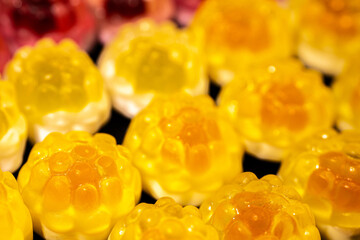 Red and yellow gummy candies resembling raspberries and pineapples are arranged on a black background. The candies have a shiny, translucent texture.
