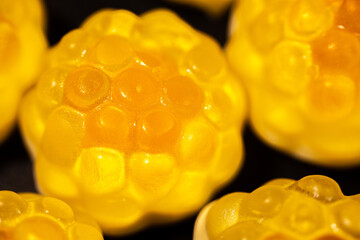 An extreme close-up of a transparent, yellow gummy candy with a bumpy, textured surface resembling a fruit or honeycomb shape. The shiny details stand out against a dark background.
