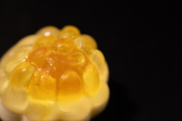 An extreme close-up of a transparent, yellow gummy candy with a bumpy, textured surface resembling a fruit or honeycomb shape. The shiny details stand out against a dark background.
