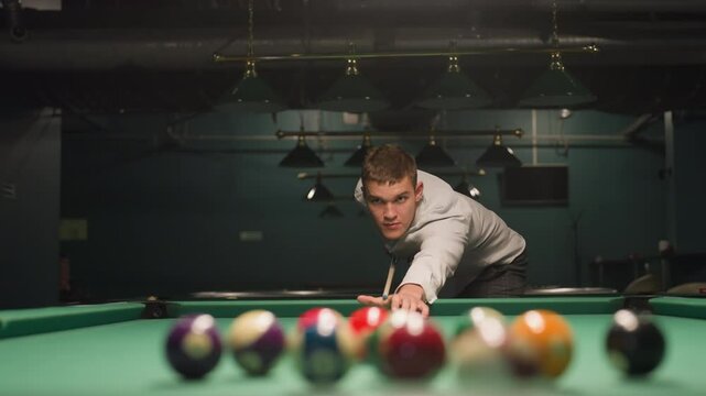 Close-up of billiard player in white shirt and grey trousers gripping cue stick with intense focus. Green pool table with scattered billiard balls. Overhead lights cast dramatic shadows
