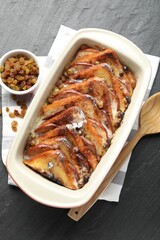Freshly baked bread pudding in baking dish, raisins and wooden spoon on dark textured table, top view