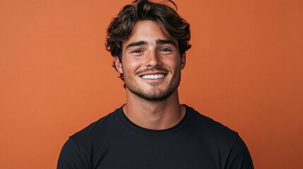 Obraz premium young stylish man in a black t-shirt smiling and standing in front of an orange background, portrait photography of a confident male model with a trendy outfit and charming expression in a studio sett