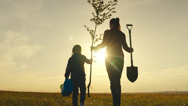 A woman and a child are planting a tree in a field. The woman is holding a shovel and the child is holding a tree. The sky is orange and the sun is setting