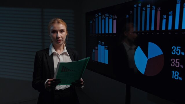 Young woman broker standing in office boardroom, analyzing financial data infographics on big display, holding folder with documents, checking information.