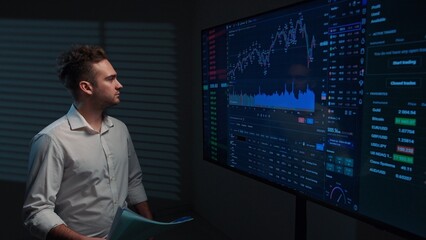 Fototapeta premium Young man broker in office boardroom analyzing financial data infographics on big display, reading report documents, looking and smiling positive.