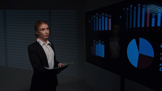Young woman broker standing in office boardroom, analyzing financial data infographics on big display, holding folder with documents, checking information.
