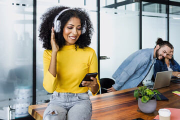 young latin woman with afro or curly hair listening music with headphones and holding mobile phone at office in Mexico Latin America, african american female in workplace