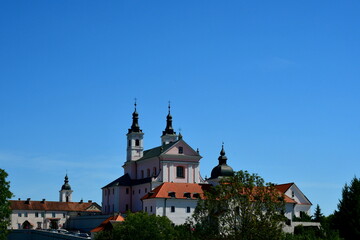 A view of a well maintained monastery or chapel located at the top of a tall hill and being surrounded with numerous trees and shrubs seen on a cloudless summer day in Poland during a hike