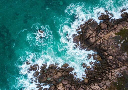 Aerial photograph of a rocky coastline with turquoise waves crashing against dark, jagged rocks, highlighting the dynamic movement and texture.

