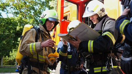 Male firefighters in protective uniforms discuss rescue plan looking at paper instructions near...