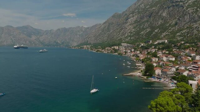 Drone view flying past the elongated roof of a church on the coastline of the city of Dobrota, along the European town of Kotor at the foot of the mountain in Kotor Bay, Montenegro, with boats and