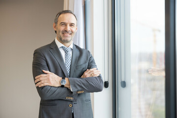 Fototapeta premium The image presents a businessman with arms crossed, radiating confidence and assurance while gazing out the window in an office.