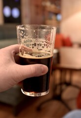 An unfinished transparent glass mug with dark beer and foam in the hand of an unknown person