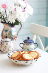 Ceramic dish with cheesecakes on a white kitchen table. There is a jug of flowers and a teapot in the background. Selective focus.