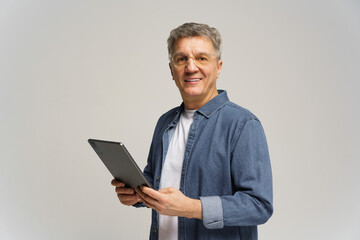 Senior man holds tablet and smiles warmly while standing against plain background in casual attire