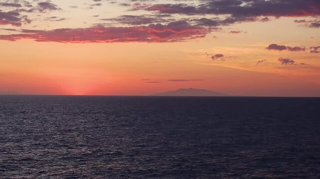 Deep rich colors of the sunset cast a brilliant glow onto the overhead clouds with a hazy island barely visible in the far distance over the Tyrrhenian Sea.