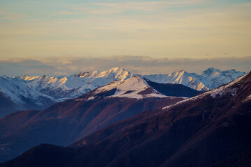 Snow-dusted mountain peaks are illuminated by warm light of sunset near Piani di Bobbio in Italy, creating striking contrast between rugged terrain and the golden sky
