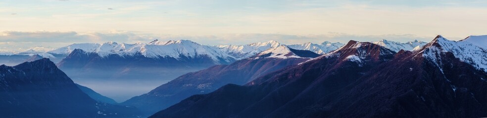 Misty valley lies beneath snow-capped mountain peaks at dusk near Piani di Bobbio in Italy, with soft light creating serene and tranquil alpine landscape