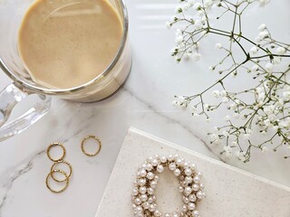 Elevated flat lay with coffee, delicate jewelry, and white flowers against a marble background. 