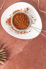 Top view of a metal sieve filled with cocoa powder on a white plate, placed on a brown surface, with some scattered cocoa powder around