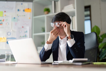 Elderly businessman showing signs of stress while working on a laptop in a modern office setting.