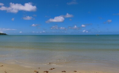 A serene stretch of beach reveals soft golden sands and gentle waves lapping the shore under a bright blue sky with scattered clouds. A small boat glides peacefully across the water