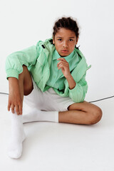 Fashionforward young boy in green jacket and white shorts sitting on white floor with trendy white...