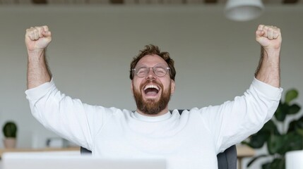 Naklejka premium Excited bearded man raising his fists in celebration while sitting at his desk in a bright home office.