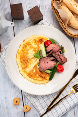 Omelet with roast beef, tomatoes and lettuce on a white plate on a wooden table. A hearty breakfast of eggs and meat, top view