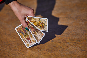 A hand holds three tarot cards with intricate illustrations against a textured wooden surface; the...