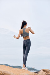 Obraz premium Fit female athlete in gray activewear stretching on a rocky beach, showcasing strength, determination, and a healthy lifestyle against a soft sky background