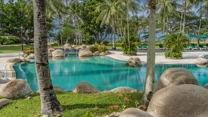 Aquamarine swimming pool in the seaside park. Picturesque boulders around the perimeter. A row of deck chairs. Green grass on lawns, palm trees. Sun umbrellas by the ocean in the distance. Malaysia. 