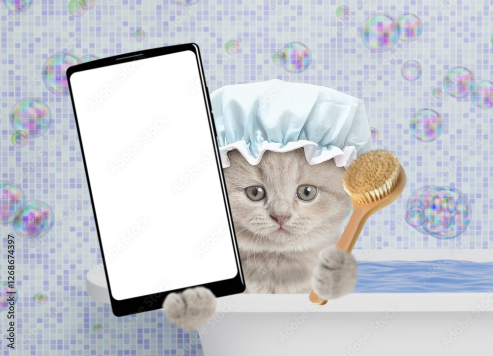 Poster cute kitten wearing showert cap takes the bath at home and shows big smartphone with white blank scr